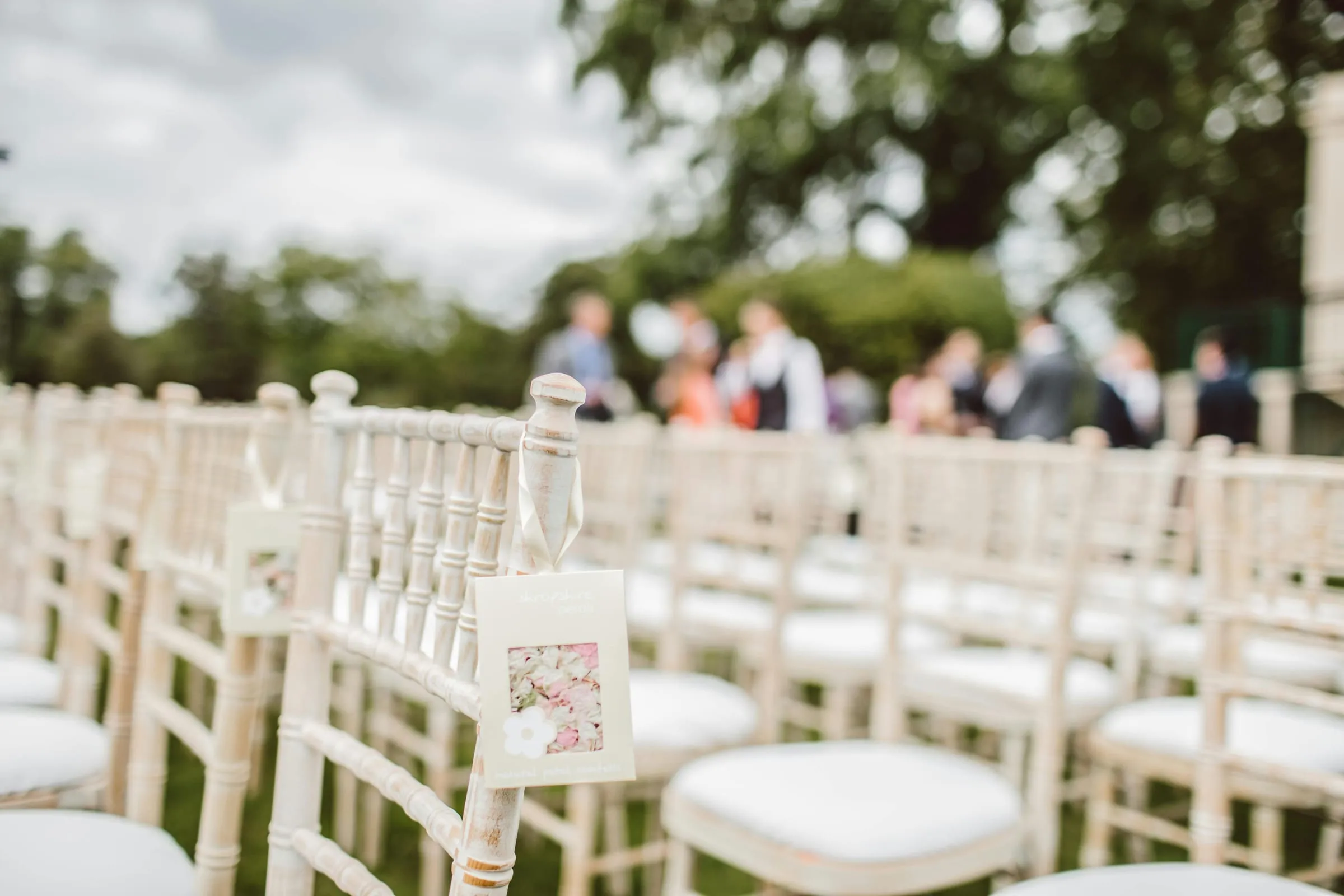 Rows of white ceremony chairs on a lawn, a packet of petal confetti tied to the end of an aisle, with guests gathering behind.