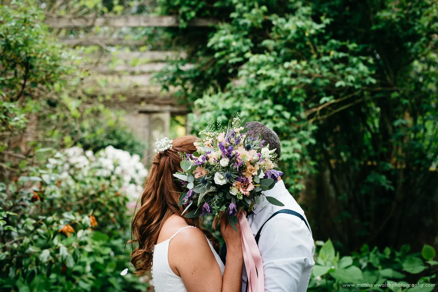 A couple kissing behind a bouquet of wildflowers in a garden.