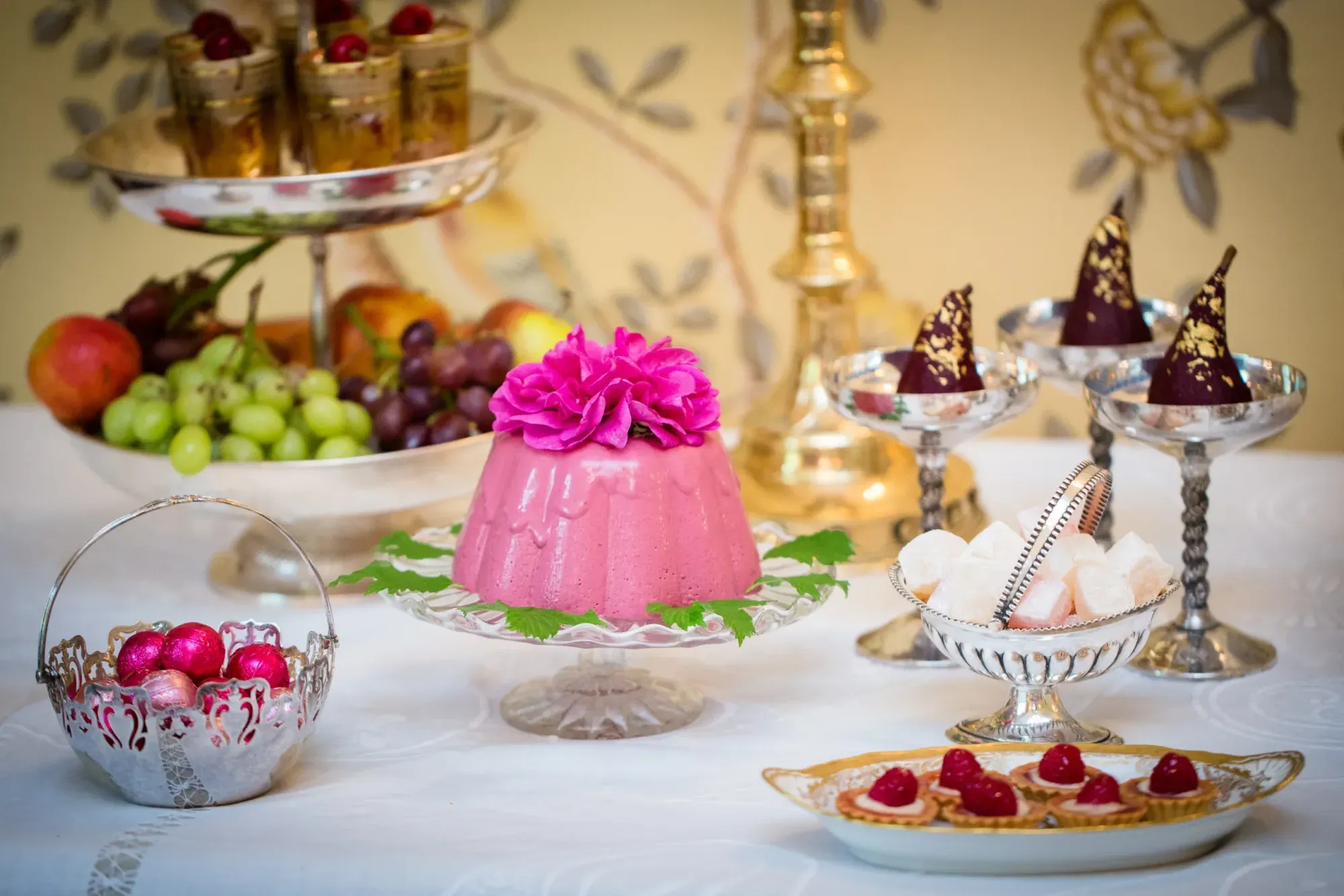 A wedding dessert table with a pink jelly, chocolate-dipped pears and fruit on silver stands.