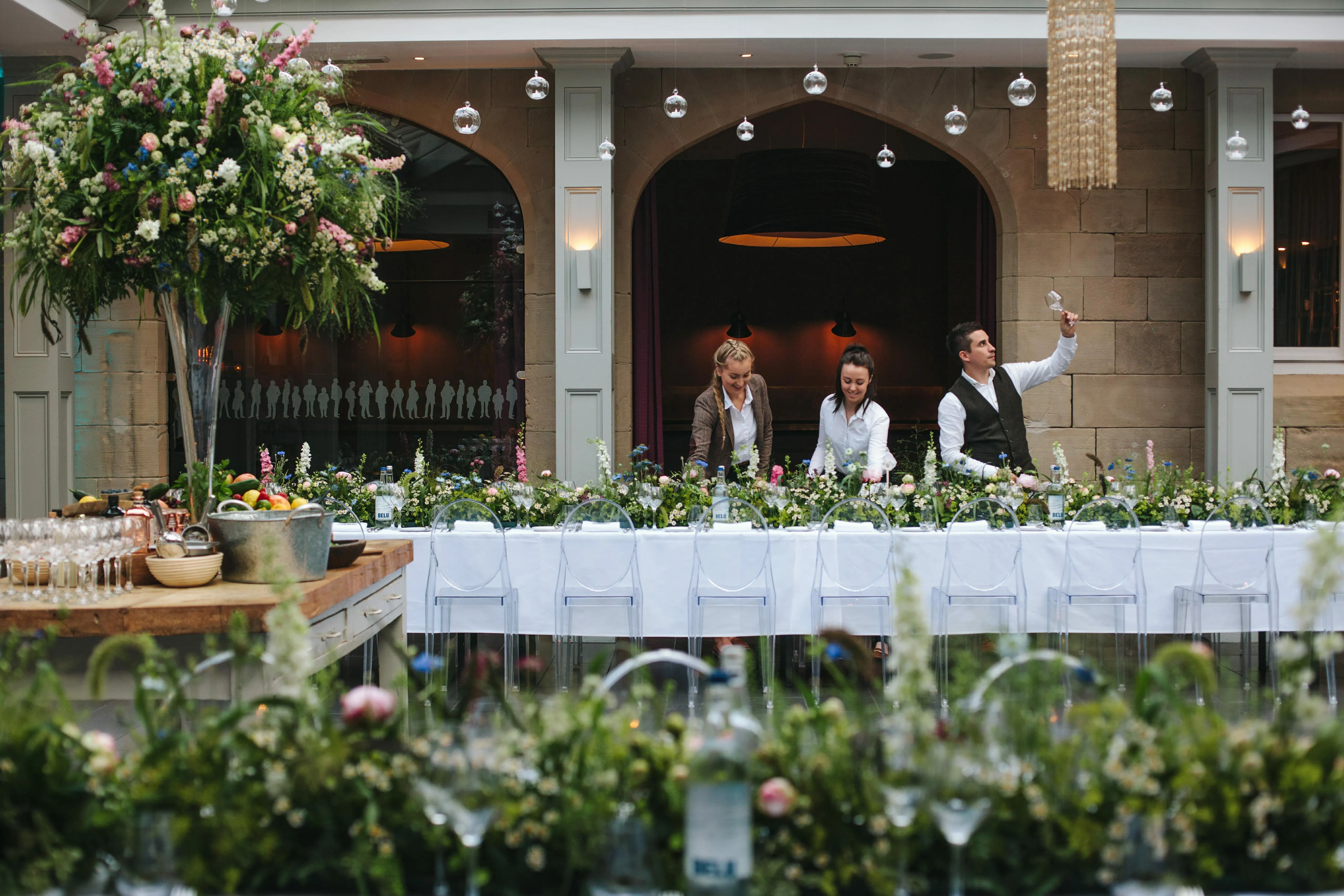 Three members of venue staff dressing a long banquet table before guests arrive, laying out glassware along a wildflower runner.