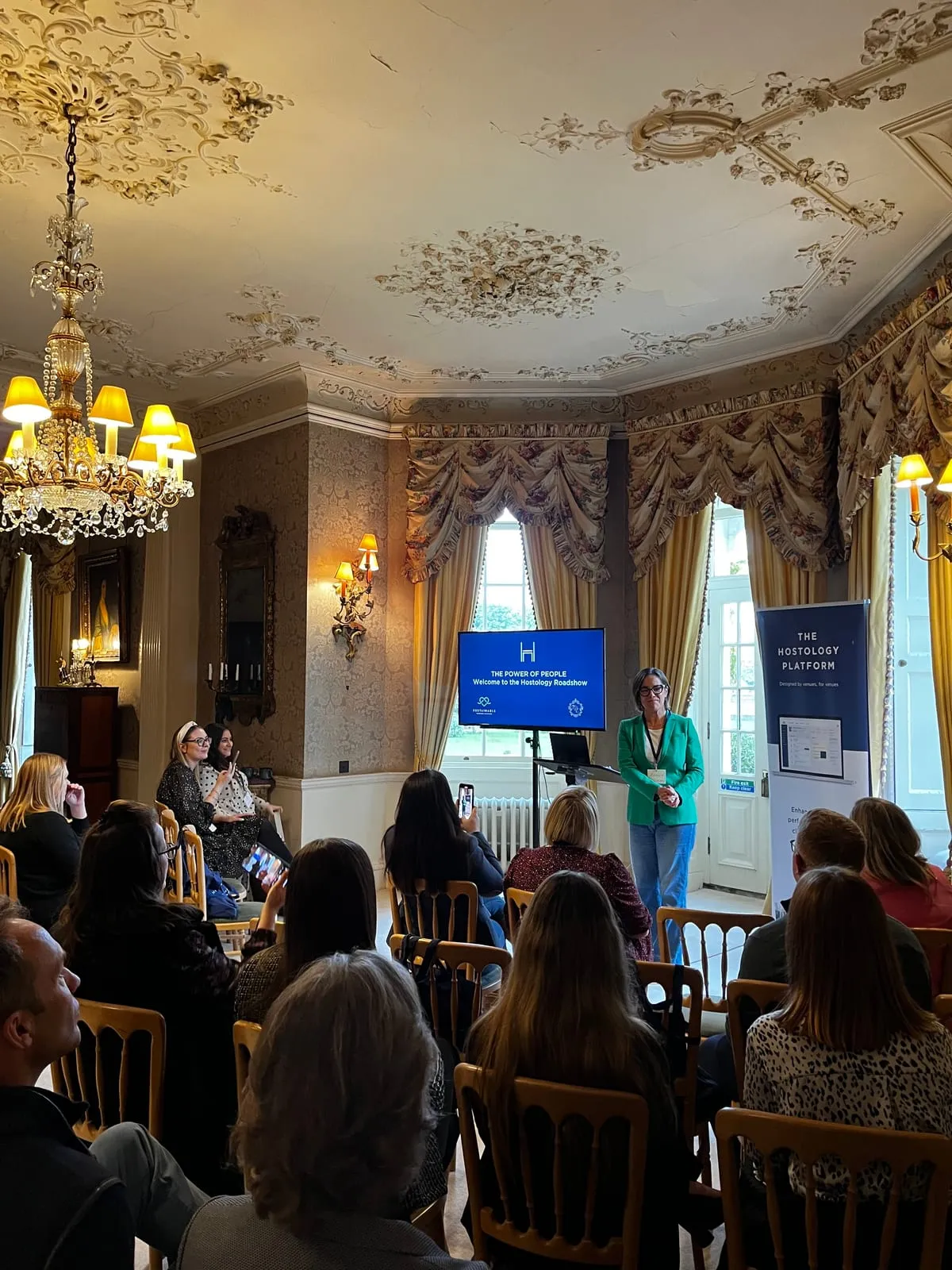 A speaker welcoming venue owners to the Hostology Roadshow, in a drawing room with an ornate plaster ceiling and a chandelier.