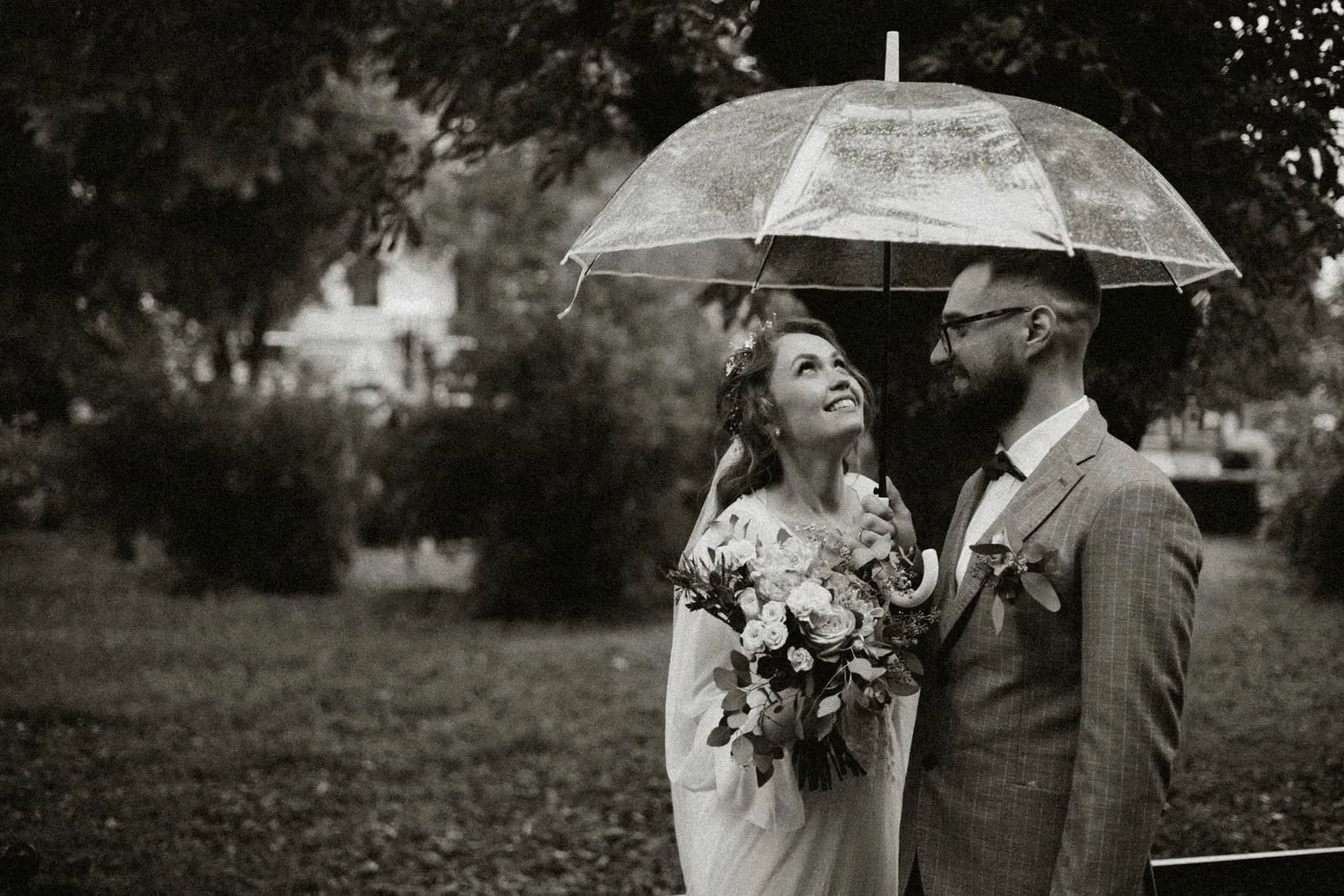 A bride and groom sharing a clear umbrella in the rain, in black and white.