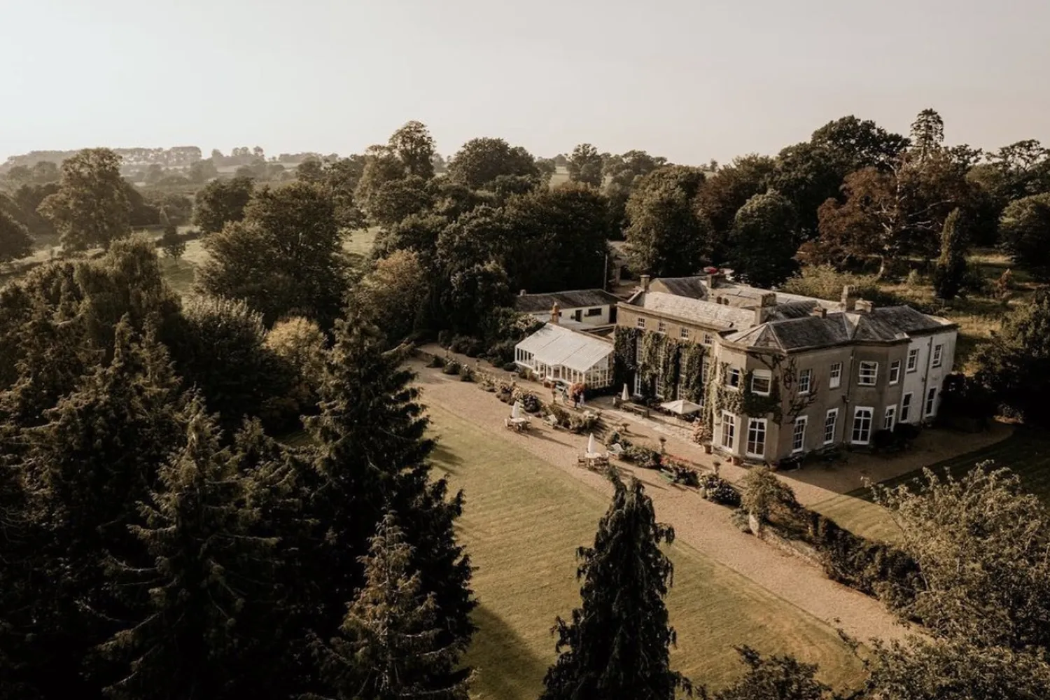 Pennard House from the air at golden hour: the house with its conservatory and a marquee on the lawn, ringed by conifers.