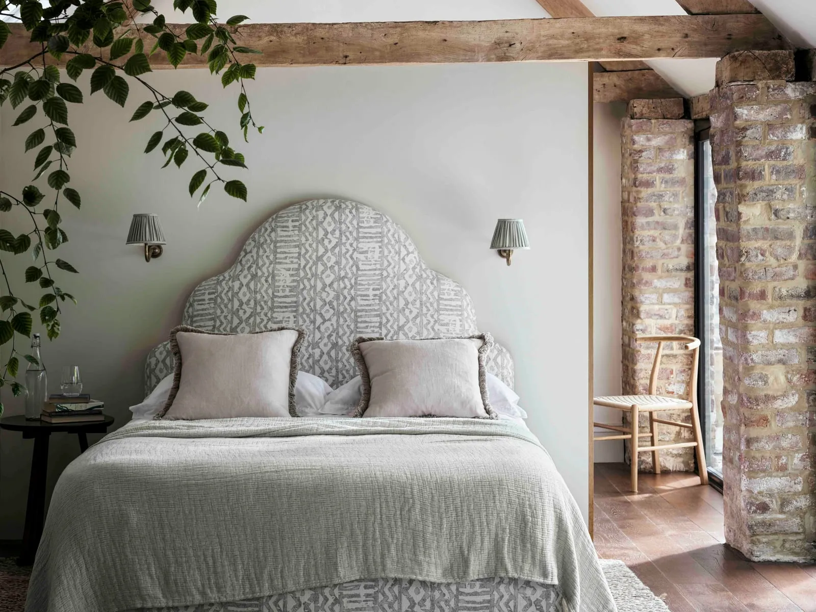 A bedroom under oak beams, with a patterned headboard, linen bedding and a chair by a doorway framed in old brick.