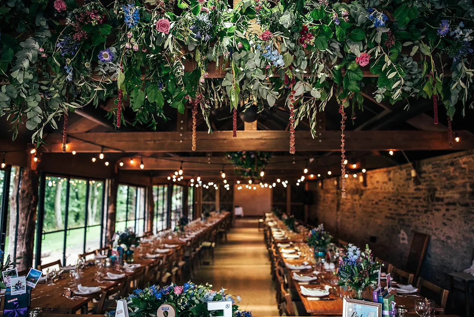 A barn laid for a wedding breakfast, with long tables, candles and garlands of flowers and greenery hanging from the beams.