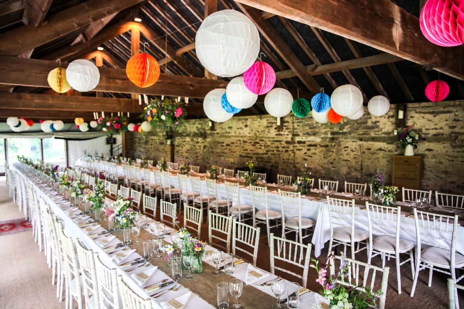 Long tables laid for a wedding breakfast in a stone barn, hung with paper lanterns.